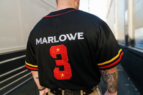 A man viewed from behind wearing a black baseball-style jersey with 'MARLOWE' in white letters and a large red number 3 with gold stars on the back. The jersey has red and yellow sleeve stripes.