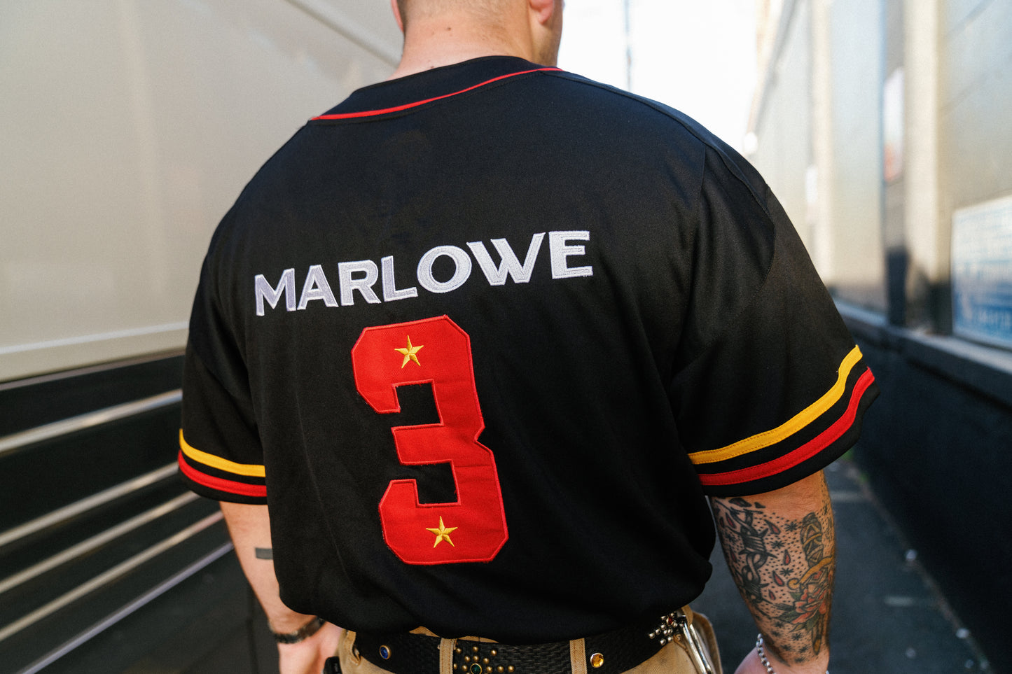 A man viewed from behind wearing a black baseball-style jersey with 'MARLOWE' in white letters and a large red number 3 with gold stars on the back. The jersey has red and yellow sleeve stripes.